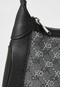 Sac à main noir avec une finition texturée, fermeture éclair et un panneau transparent orné de sequins argentés dans un motif floral.
