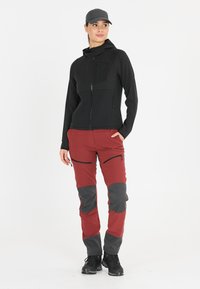 Sweat à capuche zippé noir avec un panneau texturé, associé à un pantalon rouge et gris avec zips et des empiècements contrastants aux genoux. Terminé avec des chaussures noires.