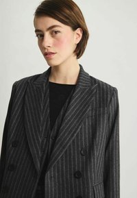 Blazer croisé en gris foncé avec des rayures blanches, fabriqué en tissu texturé, avec de grands boutons noirs et des épaules structurées.