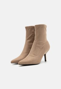 Bottes à chevilles en faux daim beige avec un bout pointu, un haut talon aiguille et une texture lisse ; design épuré avec des détails de couture minimaux.