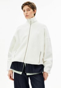 Weißes Fleece-Jacke mit hohem Kragen, Frontreißverschluss und Seitentaschen. Darüber ein blaues Jeanshemd mit lockerem Sitz und weicher Textur.