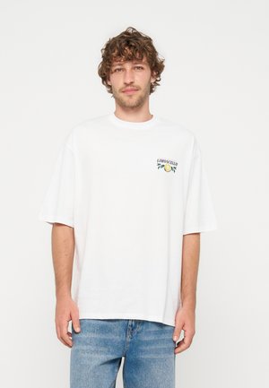 Mand med krøllet hår iført hvid oversized T-shirt med lille citrongrafik og teksten "Limoncello" på brystet, blå jeans, stående mod hvid baggrund.