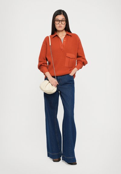 Rust-colored ribbed sweater with a pocket, paired with flared blue denim pants and a cream circular bag, featuring a chain strap.