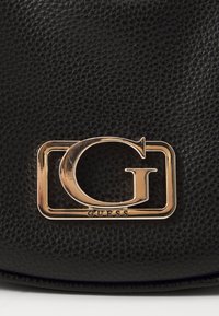 Superficie in pelle nera a forma di ciottoli con un logo Guess metallico dorato che presenta una grande "G" all'interno di una cornice rettangolare.