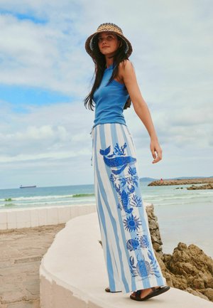 Woman wearing a blue sleeveless top, wide striped pants with marine life prints, and a straw hat, standing near a seaside with rocks and waves.