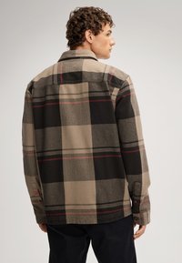 Koszula w kratę w odcieniach brązu, czerni i czerwieni. Posiada kołnierzyk, długie rękawy, zapięcie na guziki oraz teksturowaną tkaninę.