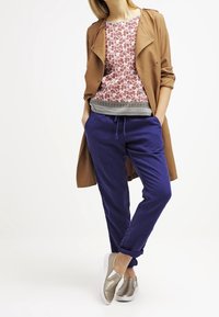 Bruine trenchcoat, bloemenprint roze top met zwarte accenten, blauwe joggingsbroek en metallic instapschoenen. Gladde texturen en levendige kleuren.