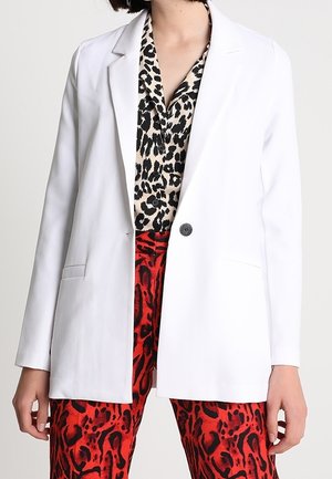 Blazer blanc à un bouton sur une blouse imprimé léopard noir et beige avec un pantalon imprimé animalier rouge et noir.