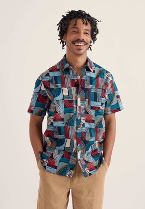 Man die lacht met handen in zakken, gekleed in een kort mouw overhemd met een kleurrijk abstract geometrisch patroon.