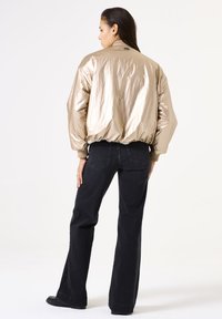 Veste bomber dorée métallique avec des poignets et un col côtelés, associée à un jean noir taille haute, vue de dos.