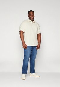 Ανοιχτό μπεζ πόλο πουκάμισο με γιακά, συνδυασμένο με μπλε τζιν. Το μοντέλο στέκεται με λευκά αθλητικά παπούτσια μπροστά σε απλό φόντο.