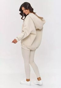 Przesadnie duża beżowa bluza z kapturem z kangurzą kieszenią, zestawiona z dopasowanymi beżowymi legginsami i białymi butami sportowymi, prezentująca miękki, teksturowany materiał.
