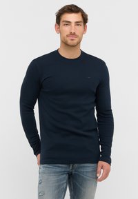 Marineblaues T-Shirt mit langen Ärmeln aus weicher Baumwolle, mit einem runden Ausschnitt und dezentem Logodetail auf der Brust, kombiniert mit hellen, lässig ausgefransten Jeans.