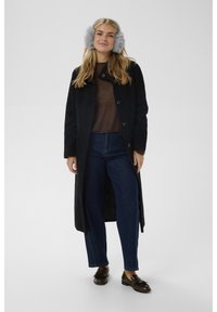 Blonde woman wearing gray earmuffs, long black coat, brown sweater, blue jeans, and brown loafers standing against plain background.