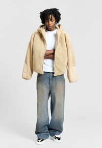 Beige fleecejacka med huva, dragkedjestängning och vida ärmar. Matchad med blekta blå jeans och sneakers med svart-vitt mönster.