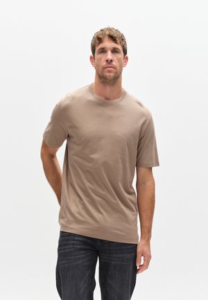 Man met lichtbruin haar, gekleed in een effen beige t-shirt en donkere spijkerbroek, staand tegen een witte achtergrond met één hand achter zijn rug.