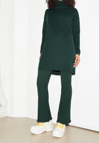 Osoba v tmavě zeleném dlouhém svetru a odpovídajících kalhotách, s bílými chunky teniskami se žlutými tkaničkami, stojící uvnitř.