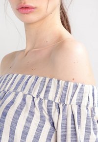 Haut dévoilant les épaules avec un col froncé, présentant des rayures horizontales bleues et blanches sur un tissu texturé. Teint de peau lisse visible à proximité.