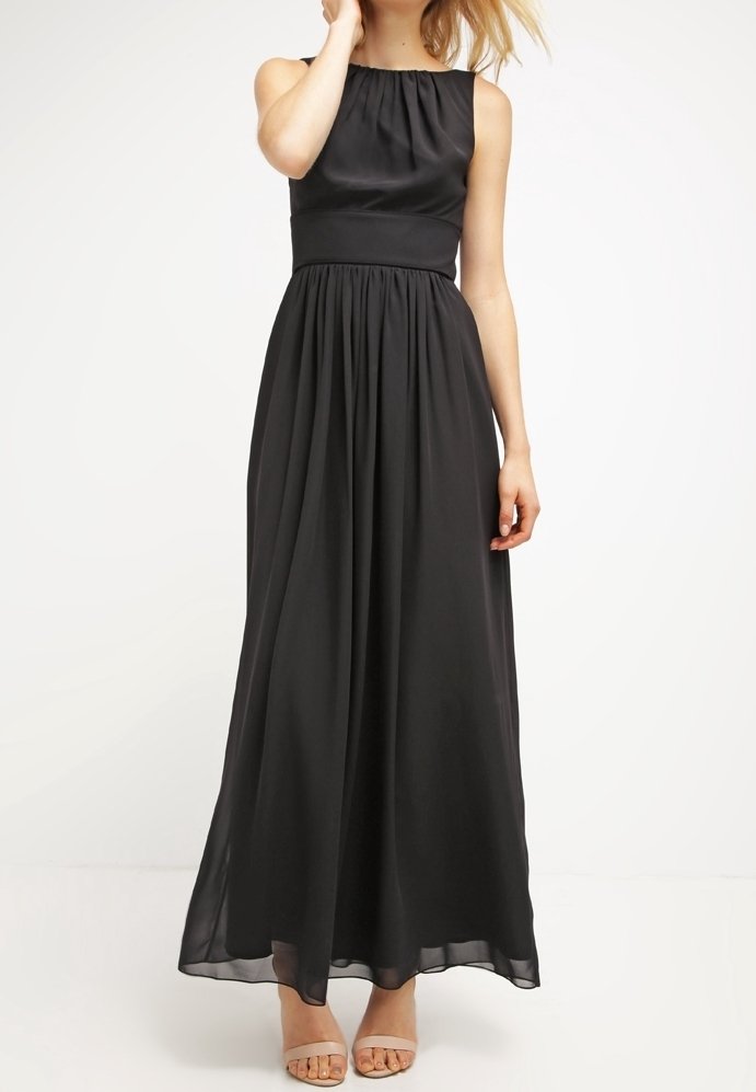 Femme portant une robe noire sans manches longue jusqu’au sol avec taille et encolure froncées, associée à des sandales ouvertes couleur nude, debout sur fond blanc.