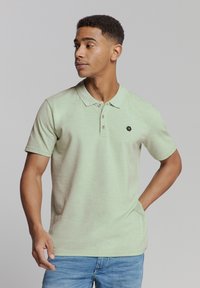 Lichtgroene polo met een kraag, twee knopen en een klein zwart logo. Korte mouwen, zachte textuur en casual pasvorm. Draag het met blauwe spijkerbroeken.