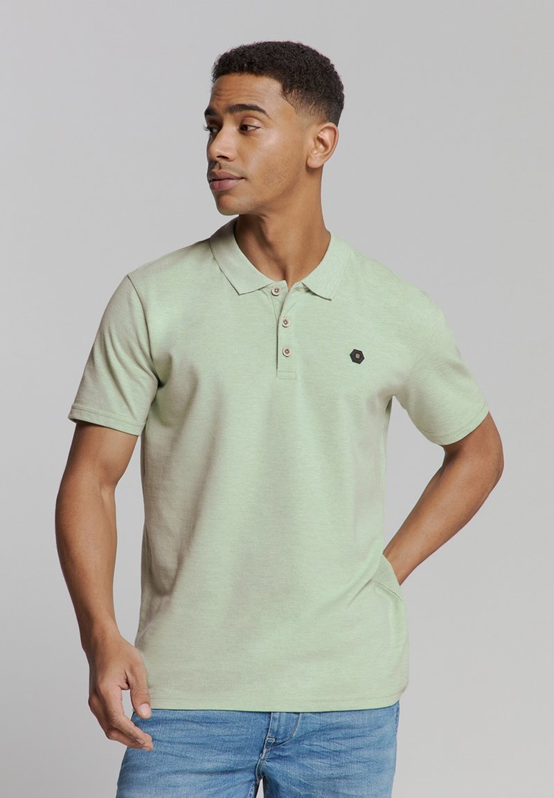 Lichtgroene polo met een kraag, twee knopen en een klein zwart logo. Korte mouwen, zachte textuur en casual pasvorm. Draag het met blauwe spijkerbroeken.