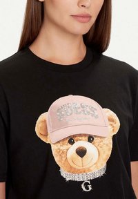 Camiseta negra con un gráfico de un oso de peluche que lleva una gorra rosa con lentejuelas. El oso tiene un collar con joyas y detalle de logo.
