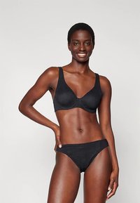 Mosolygó nő fekete melltartóban és illő alsóneműben, egyik kezét a csípőjén pihenve, egy egyszínű világos háttér előtt áll.