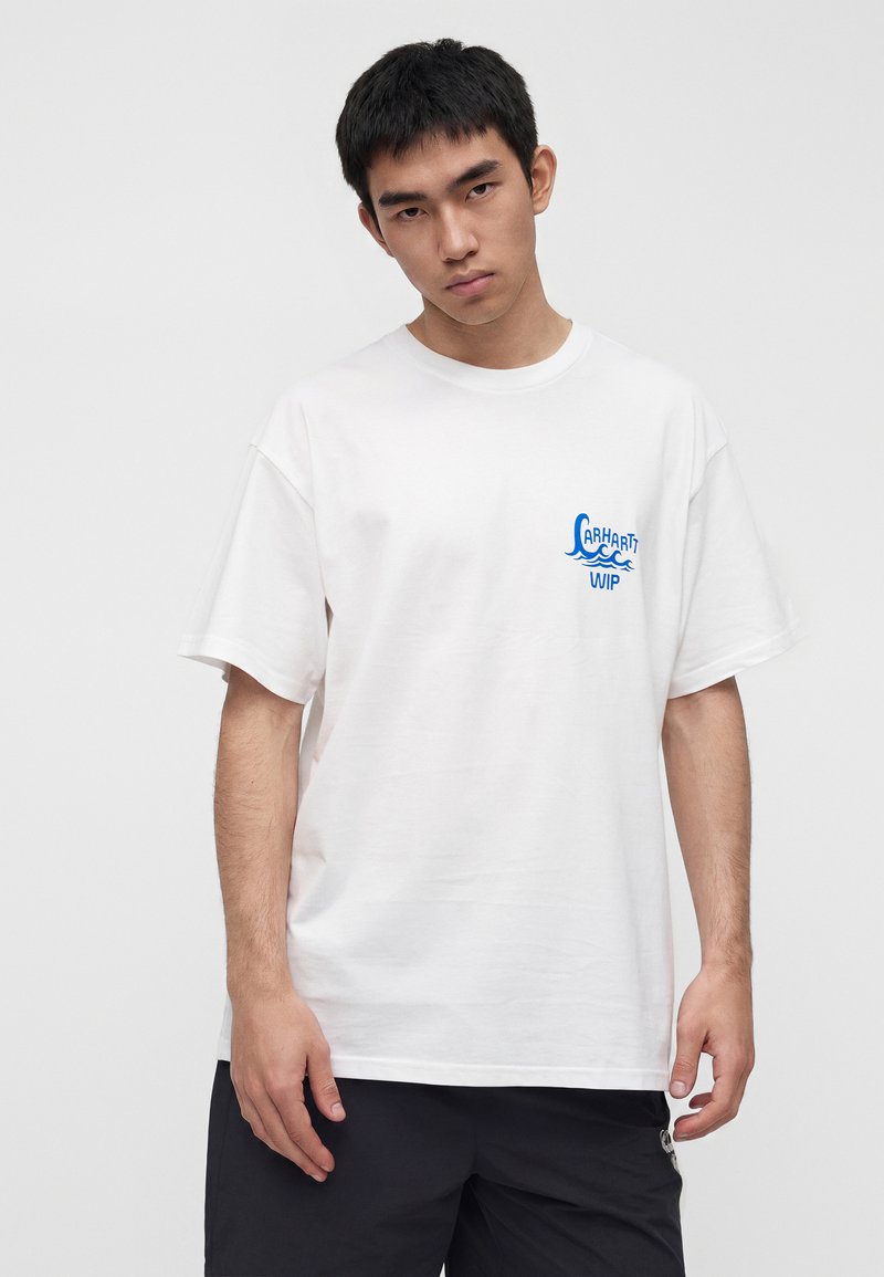 Giovane uomo con una t-shirt bianca larga con il logo blu "Carhartt WIP" sul petto, abbinata a shorts neri, in piedi davanti a uno sfondo grigio semplice.