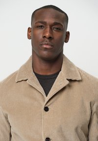 Beige corduroy jas met een kraag, voorzien van verticale ribbels, twee zwarte knopen en een gestructureerde afwerking over een donker t-shirt.
