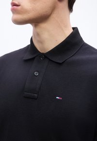 Νέος άνδρας που φοράει μια μαύρη πόλο μπλούζα με δύο κουμπιά και ένα μικρό ορθογώνιο λογότυπο σε κόκκινο, λευκό και μπλε στο στήθος.