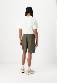 Person med lange fletninger iført en hvid t-shirt, olivengrønne cargo shorts, hvide sokker og beige sneakers, stående mod en hvid baggrund.