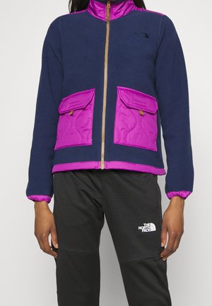 Osoba nosząca granatową kurtkę polarową z fioletowymi pikowanymi kieszeniami, kołnierzem i lamówką, w połączeniu z czarnymi spodniami z białym logo North Face.
