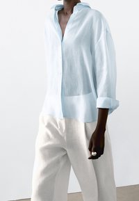 Camicia di lino azzurro chiaro con colletto, chiusura a bottoni sul davanti e maniche arrotolate, abbinata a pantaloni di lino color crema. Texture morbida, vestibilità rilassata.