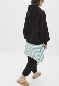 Hoodie noir surdimensionné, chemise rayée verte et blanche superposée en dessous, jogging noir et chaussures beige doublées de fourrure avec des accents de boucle.