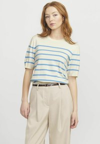 Stribet strikbluse i creme med blå striber, korte ærmer, rund hals. Parret med beige plisserede bukser og et mørkt bælte.