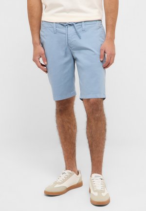 Mann trägt hellblaue Shorts mit Kordelzug und weiße Sneaker mit beigen Akzenten, steht vor einem schlichten hellen Hintergrund.