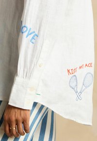 Valge linane särk, millel on sinine "LOVE" ja oranž "KISS MY ACE" tikand ning kaks tennisesaabikut. Nööpidega mansett õrna voltimise detailiga.