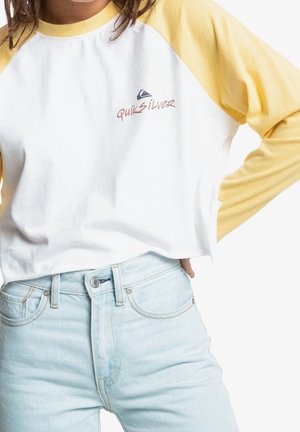 Osoba w białej koszulce Quiksilver z żółtymi rękawami, włożonej w jasnoniebieskie dżinsy, z ręką opartą na biodrze na białym tle.
