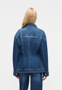 Ξανθός άνθρωπος στέκεται με την πλάτη, φορώντας μια εφαρμοστή μπλε τζιν ζακέτα και ταιριαστό μπλε τζιν παντελόνι μπροστά σε ένα απλό φόντο.