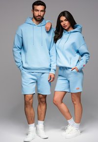 Světle modré mikiny s kapucí a odpovídající šortky, vyrobené z měkkého materiálu, s uvolněným střihem a malými logovými akcenty. Na sobě bílé tenisky.