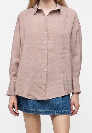 Femme portant une chemise rose pâle ample à manches longues avec des boutons et une jupe courte en denim bleu, debout devant un fond blanc uni.