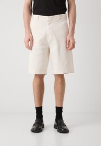 Beige Baumwollshorts mit geradem Schnitt, seitlichen Taschen und glatter Textur, kombiniert mit schwarzen Socken und Slippern.