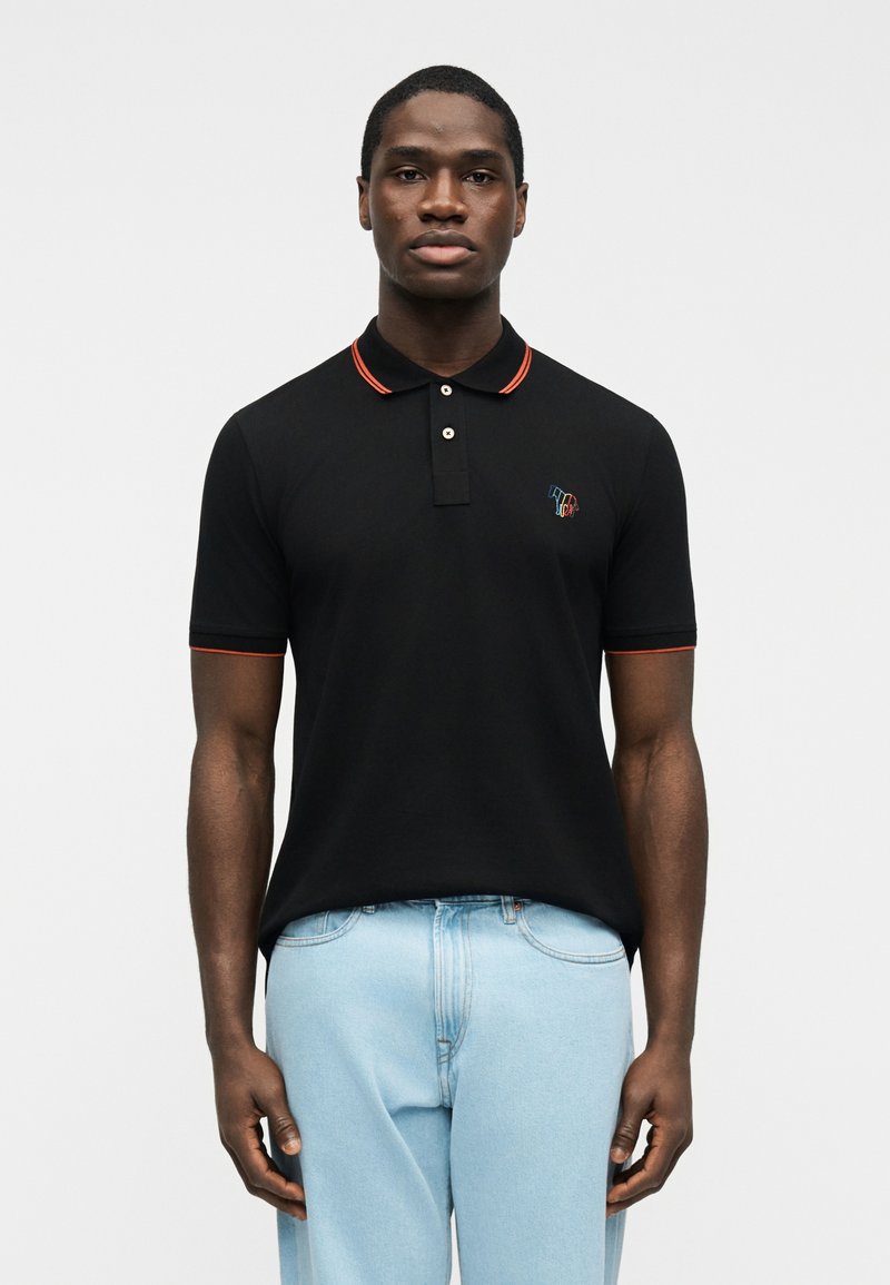 Άνδρας που στέκεται μπροστά σε απλ ό φόντο, φορώντας μαύρο πόλο με κόκκινη επένδυση και ανοιχτόχρωμο τζιν, κοιτάζοντας μπροστά με τα χέρια χαλαρά.