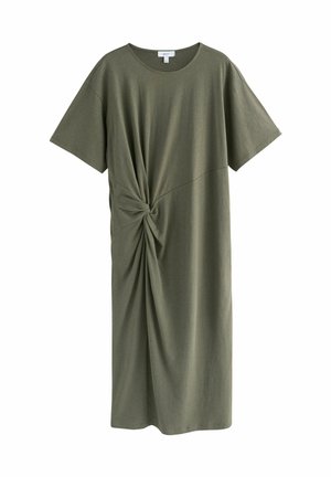 Vrouw met donker haar in een lage knot, draagt een losse groene midijurk met een split aan de achterkant, zwarte sandalen en gouden armbanden, staande bij een witte muur.