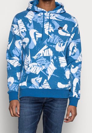 Person, die einen blauen Nike-Hoodie mit durchgehenden weißen und hellblauen Nike-Logos trägt, kombiniert mit dunkelblauen Jeans, stehend vor einem einfarbigen Hintergrund.