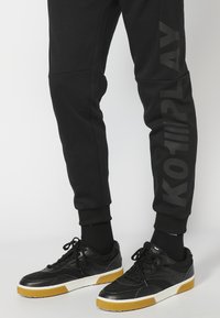 Svarte joggebukser med teksturerte sidelapper har et stort "KOTPLAY"-trykk. Kombinert med svarte sneakers og gummisåler.
