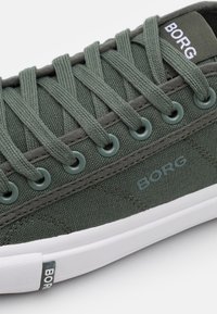 Zbliżenie zielonego płóciennego trampka sznurowanego płaskimi zielonymi sznurowadłami, białą gumową podeszwą oraz brandingiem "BORG" na języku, boku i tabliczce na podeszwie.
