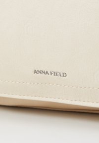 Pochette en cuir synthétique couleur crème avec logo "ANNA FIELD" en relief. Présente une texture lisse et des coutures contrastantes le long des bords.