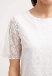 Valkoinen läpikuultava paita, jossa on monimutkaiset kirjailut kuvioineen, joissa on kiehkurat ja kukka-aiheet, lyhyet hihat ja pyöreä pääntie.