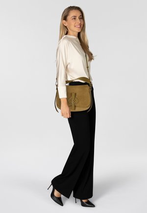 Bolso bandolera de ante verde oliva con detalle de cierre frontal, combinado con una blusa crema y pantalones negros de pierna ancha.